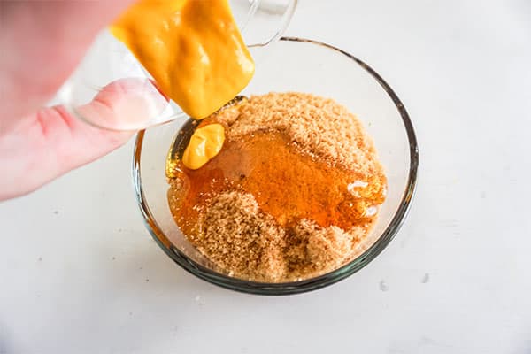 mustard being poured into ketchup, brown sugar and honey in a glass bowl on a white background