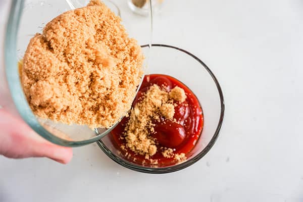 brown sugar being poured into a glass bowl of ketchup on a white background