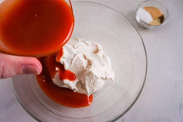 a hand pouring hot sauce into cream cheese in a glass bowl next to a glass bowl of seasonings on a white background
