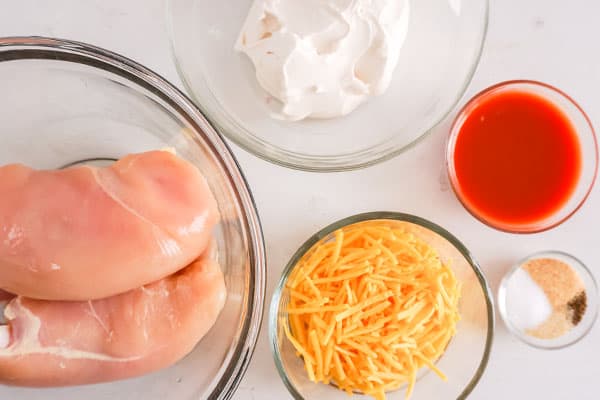 raw chicken breasts, cream cheese, hot sauce, shredded cheddar cheese, seasonings in glass bowls on a white background
