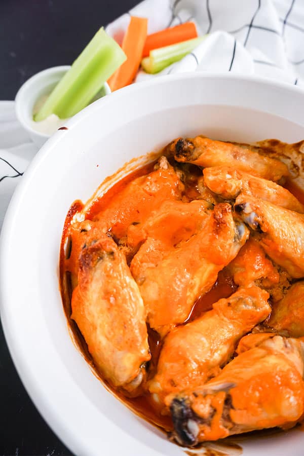 Baked Buffalo Chicken Wings in a white dish next to celery in a dip with more carrots and celery on a white cloth in the background