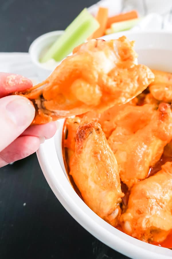 a hand holding a Baked Buffalo Chicken Wings above a white dish next to celery in a dip with more carrots and celery on a white cloth in the background
