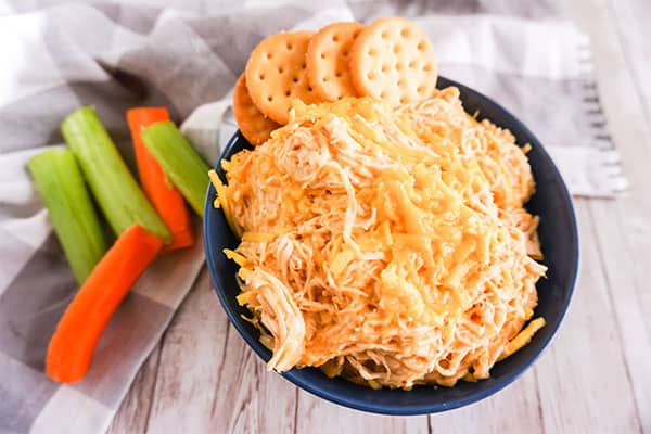 Slow Cooker Buffalo Chicken Dip in a blue bowl with some crackers next to carrot and celery sticks on a gray checkered cloth
