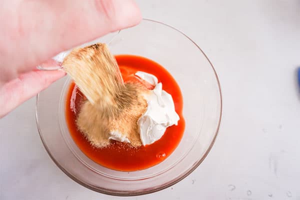 spices being poured into a glass bowl of cream cheese and hot sauce on a white background