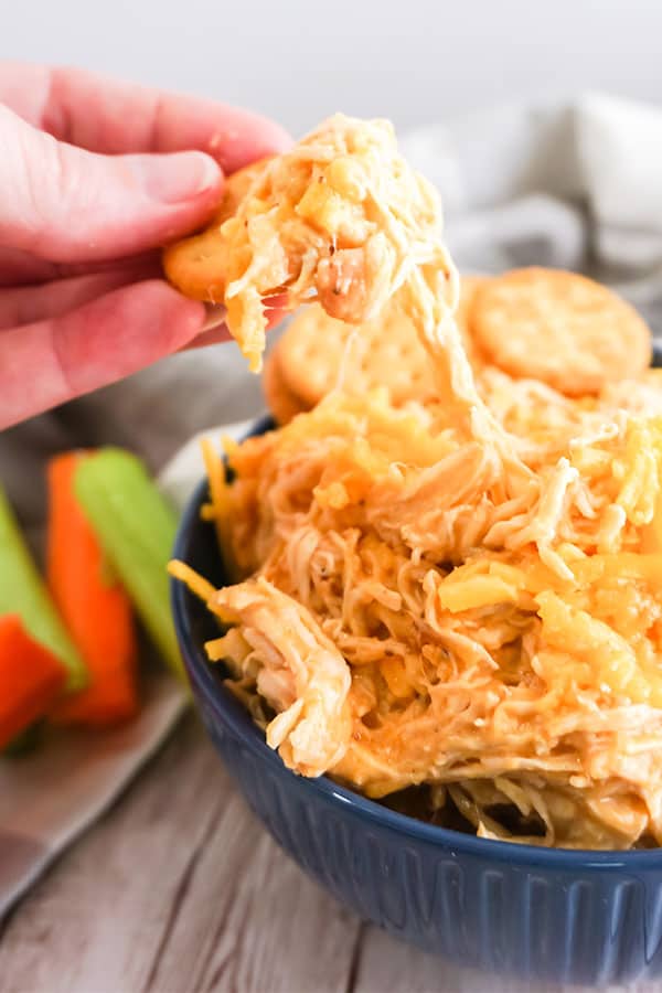 a hand holding a cracker with some dip on it with more Slow Cooker Buffalo Chicken Dip in a blue bowl 
