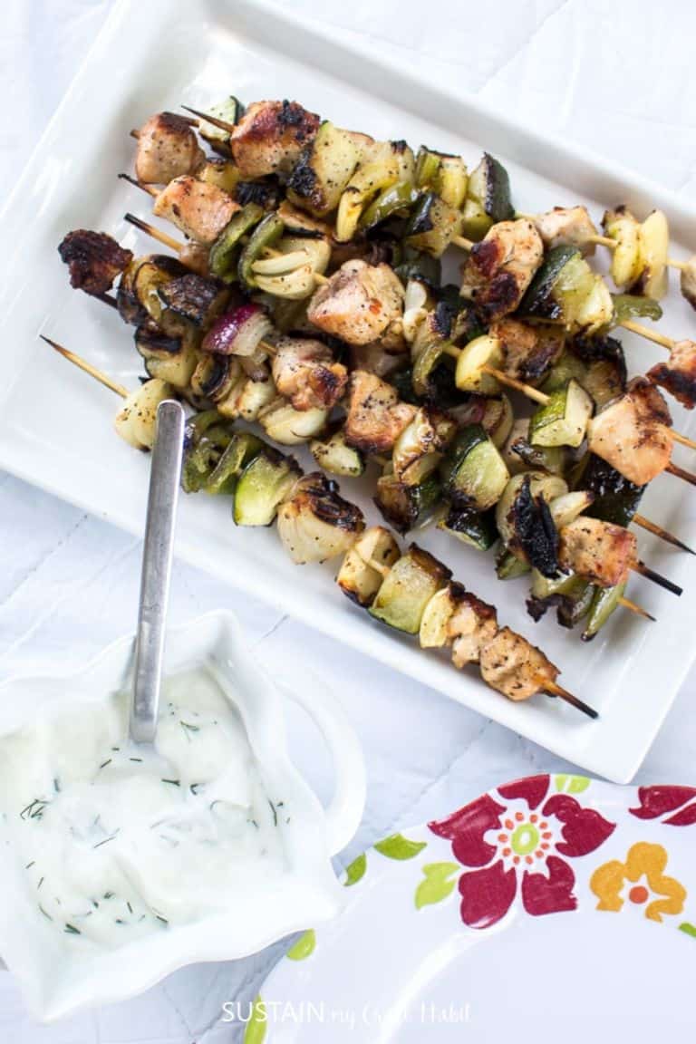 chicken shish kabobs on a white tray next to creamy dill dip in a white dish next to a flowered plate on a white background