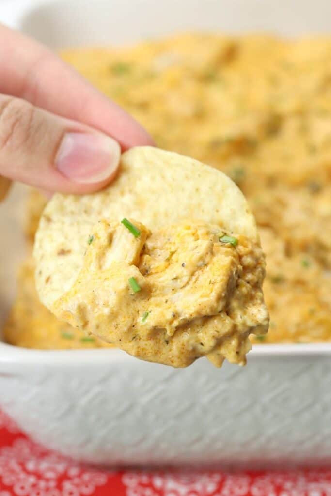 a hand holding a chip with buffalo chicken dip on it with more dip in a white dish in the background