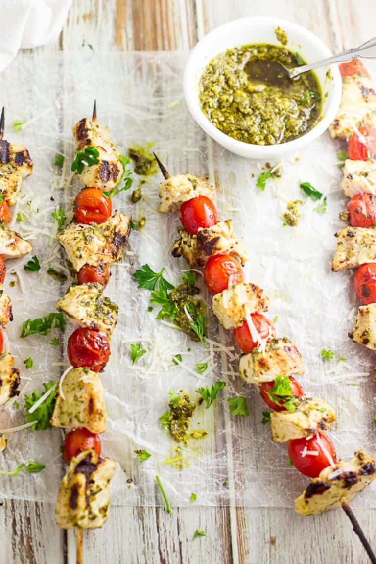 pesto chicken kabobs on parchment paper on a wood table next to a white bowl of pesto
