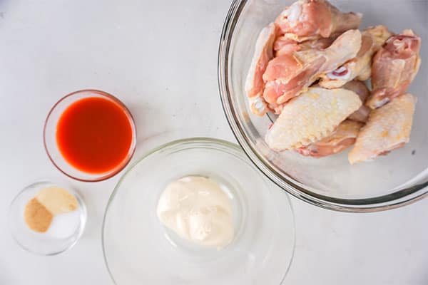 seasonings, hot sauce, miracle whip and raw chicken wings in glass bowls on a white background