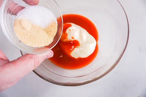 seasonings being added from a glass bowl to hot sauce and miracle whip in a glass bowl on a white background