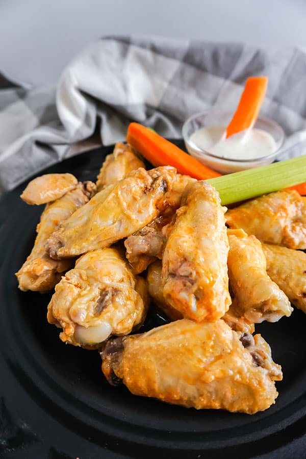 buffalo chicken wings next to carrot and celery sticks and a bowl of sauce on a black plate next to a gray cloth