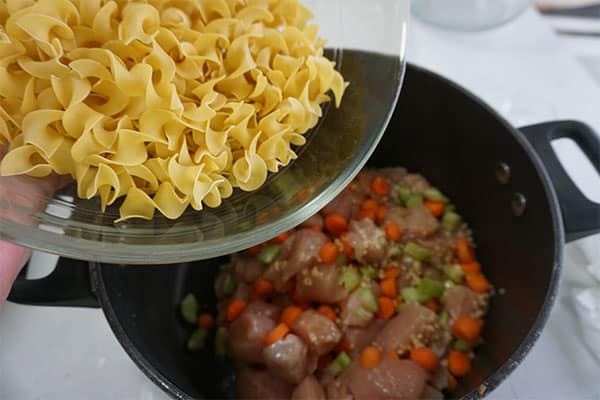egg noodles being poured from a glass measuring cup into cubed raw chicken and veggies in a pot