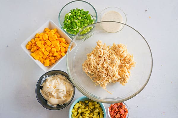 bowls of cubed cheese, chopped chives, mayonnaise, shredded chicken, bacon pieces, chopped pickles on a white background
