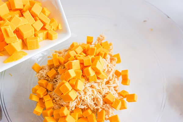 cubed cheese being added to a glass bowl of shredded chicken, mayonnaise, celery and seasoning