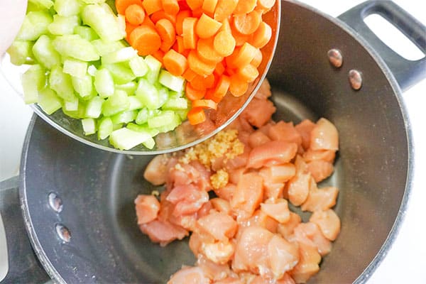 diced carrots and celery being poured into seasoning and cubed raw chicken in a pot
