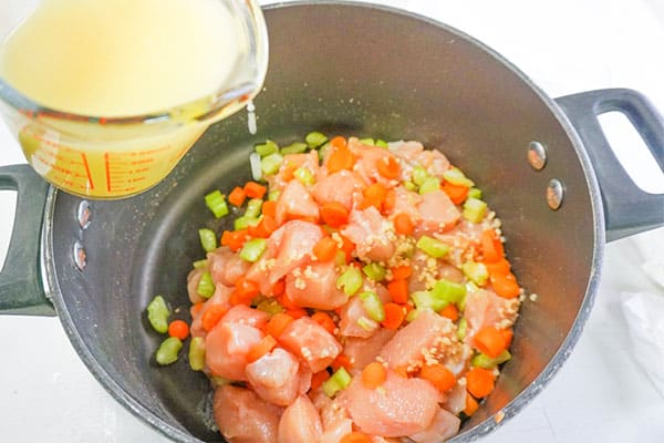 broth being poured from a glass measuring cup into cubed raw chicken and veggies in a pot