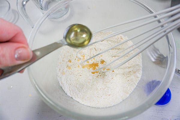 a teaspoon of vegetable oil being added to dry ingredients for dumplings being combined in a glass bowl with a whisk
