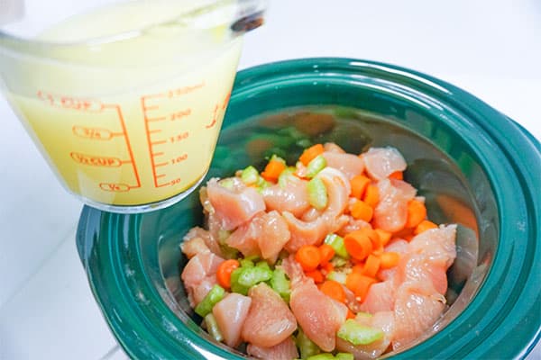 chicken broth in a glass measuring cup about to be added to raw cubed chicken and diced carrots and celery in a slow cooker