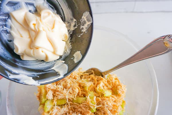 mayonnaise being poured into a glass bowl of seasoned salt, shredded chicken and chopped celery
