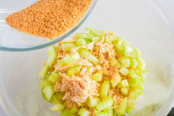 seasoned salt being poured from a glass bowl into a glass bowl of shredded chicken and chopped celery