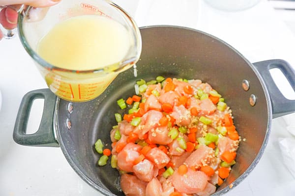 broth being poured into a pot of raw cut up chicken, celery and carrots and garlic
