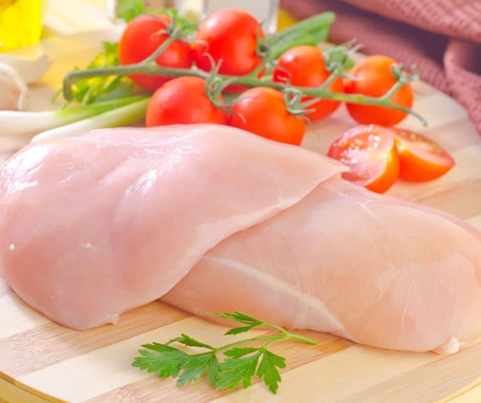 two chicken breasts on a wooden board next to tomatoes on a vine