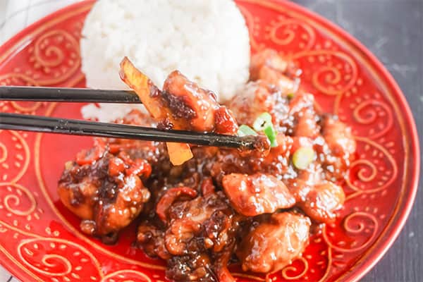 chopsticks holding some Slow Cooker General Tso's Chicken above more of the food next to some rice on a red plate