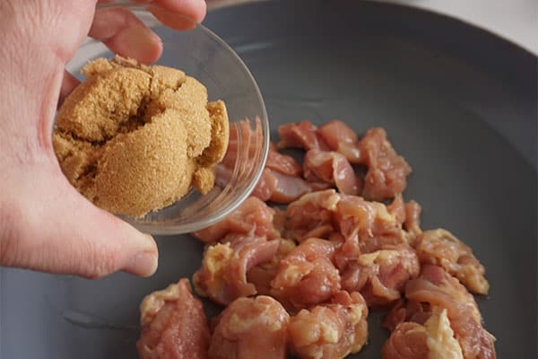 a hand adding brown sugar to cubed raw chicken breasts in a skillet