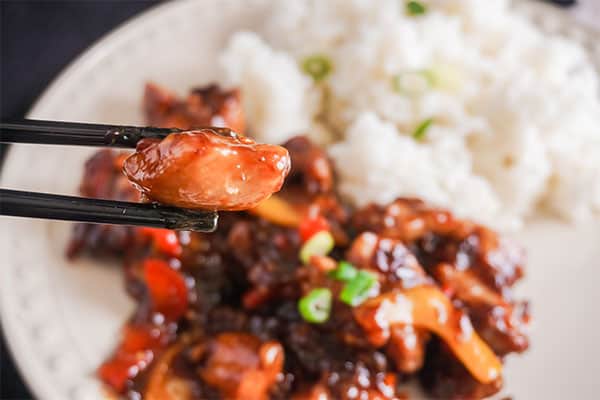black chopsticks holding a piece of Spicy General Tso’s Chicken above a plate of more of the chicken and rice