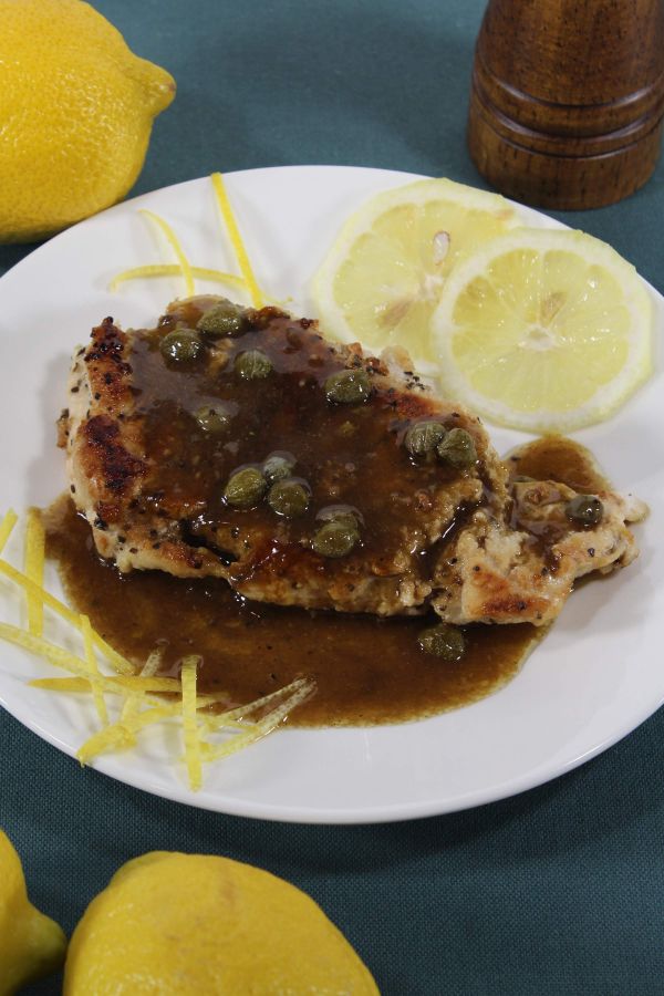 Lemon Chicken Piccata  next to lemon slices and lemon zest on a white plate on a dark blue cloth next to some lemons and a salt or pepper shaker