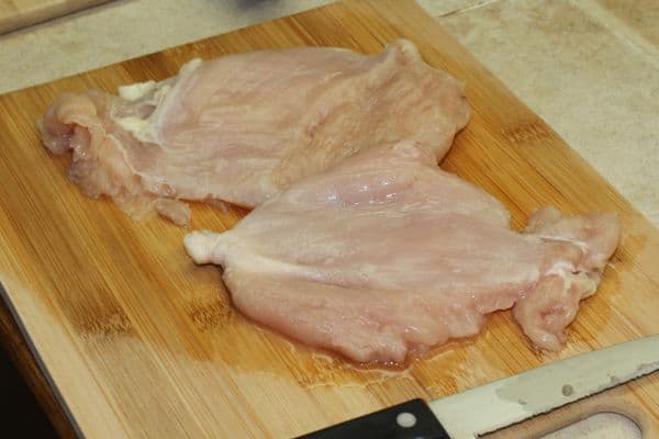flattened raw chicken breasts on a wooden cutting board next to a knife