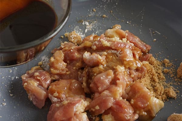 soy sauce in a glass bowl above a pan of diced chicken, cornstarch and brown sugar being cooked in sesame oil