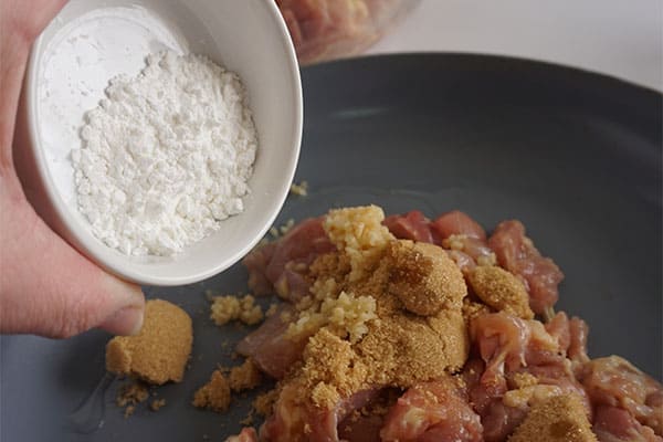 a hand about to pour cornstarch from a white bowl into a pan of diced chicken and brown sugar being cooked in sesame oil