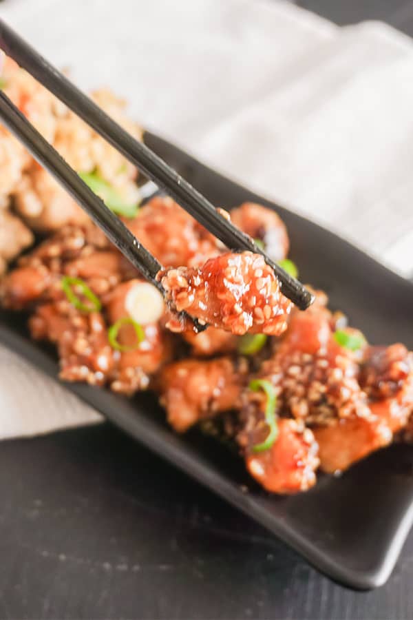 closeup of chopsticks holding a piece of chicken with more Take-Out Style Sesame Chicken on a black plate next to brown rice , on a white cloth on a brown table