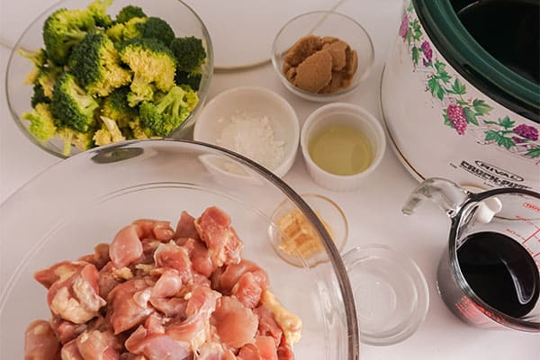 glass bowls and white containers of cubed raw chicken breasts, broccoli, brown sugar, rice wine vinegar, sesame oil, minced garlic, cornstarch, and soy sauce, all on a withe counter next to a slow cooker