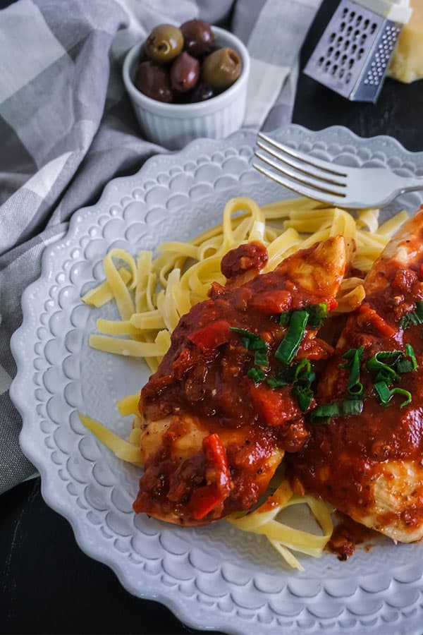 Homemade Chicken Cacciatore next to a fork on a white plate on a table next to a gray and white checkered cloth, a bowl of olives, a mini cheese grater, and a block of cheese