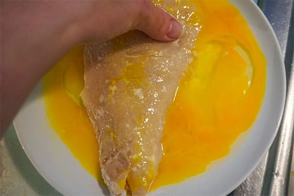 a hand dipping a raw chicken breast in egg