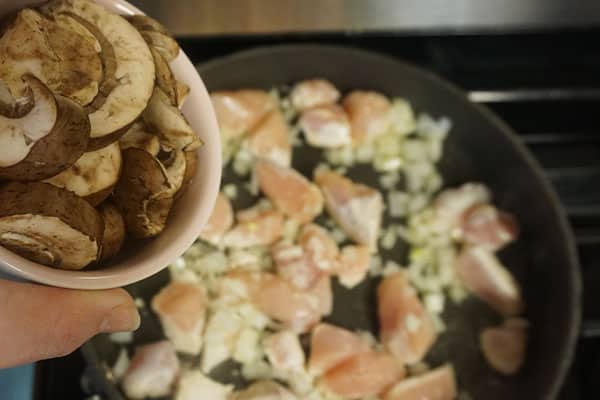 mushrooms being added to diced raw chicken breasts, onions and seasonings cooking in a skillet