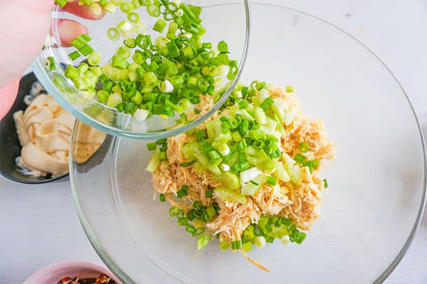 chopped chives being added to shredded chicken and sliced celery in a glass bowl next to a bowl of mayonnaise 