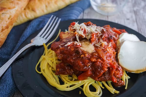 Slow Cooker Chicken Cacciatore next to a fork on a black plate on a wood table with bread, a blue cloth and a glass in the background