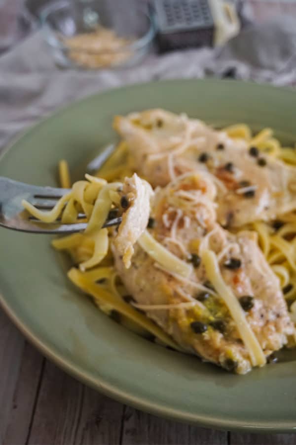 a forkful of Slow Cooker Chicken Piccata and noodles above more of the food on a green plate on a wood table with a bowl of cheese and a grater in the background