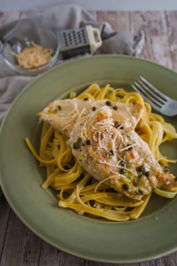 Slow Cooker Chicken Piccata and noodles next to a fork on a green plate on a wood table with a bowl of cheese and a grater in the background