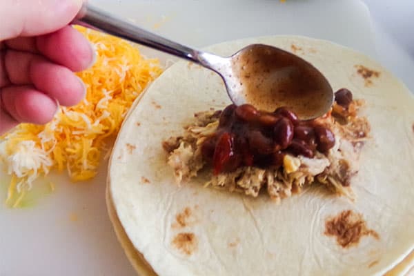 a hand using a spoon to put beans on shredded chicken on a flour tortilla with shredded cheese in the background