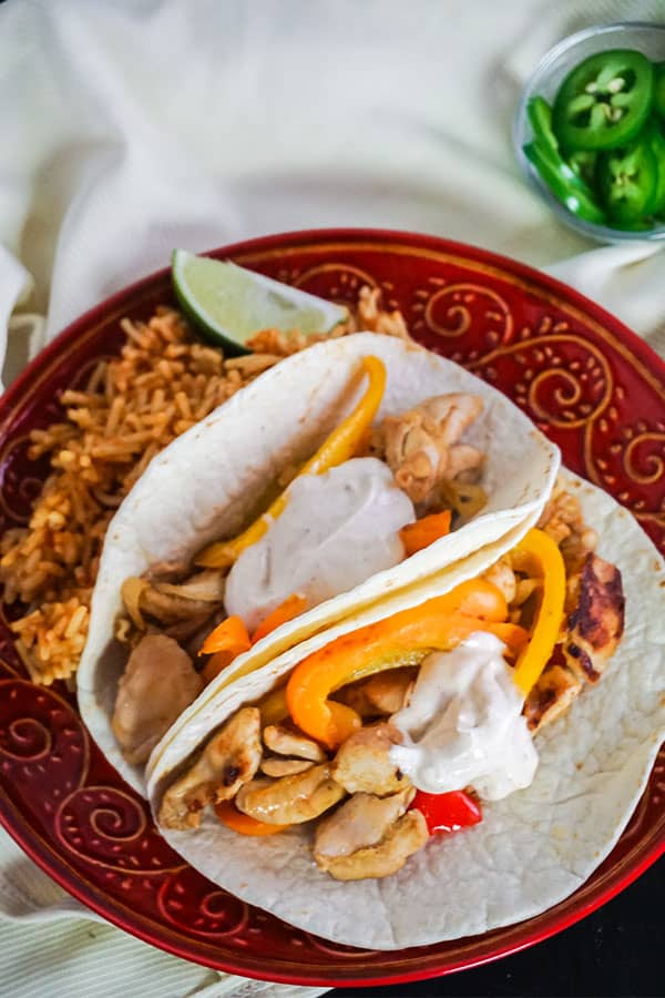 two chicken fajitas next to rice and a slice of lime on a red plate on a white cloth with a glass bowl of jalapeno slices in the background