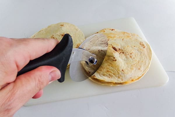 a hand using a pizza cutter to slice a chicken quesadilla on a white cutting board