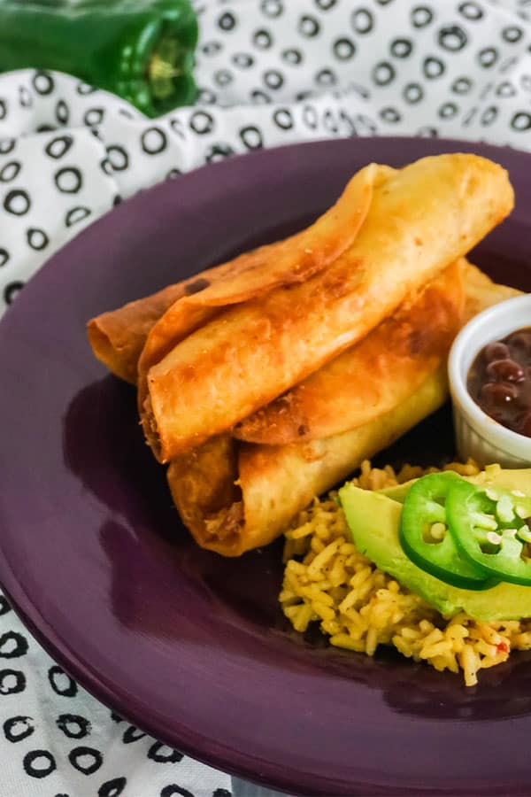 Chicken Taquitos next to rice and beans on a purple plate on a white and black cloth