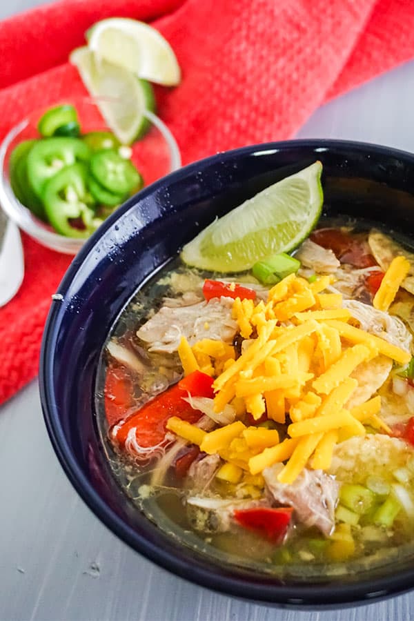 Chicken Tortilla Soup in a blue bowl on a white table next to a bowl of jalapeno slices and lime slices on a red cloth