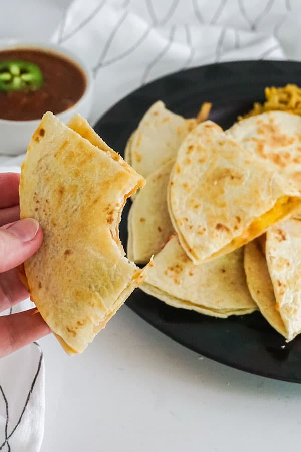 a hand holding a slice of chicken quesadilla above more sliced chicken quesadilla next to rice on a black plate on a white and black cloth next to a white bowl of salsa