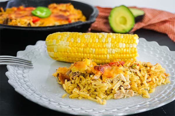 Mexican Chicken and Rice Casserole next to beans and corn on the cob on a white plate on a black table with more casserole. an avocado cut in half and a red and white checkered cloth in the background