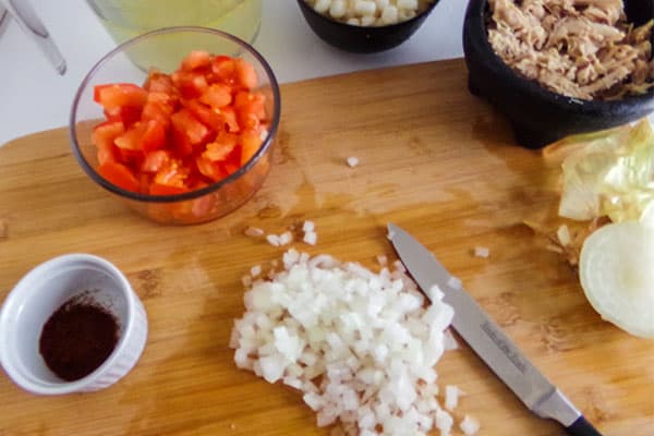chopped onion on a wooden cutting board next to a knife, and bowls of diced tomatoes, sauce and chicken